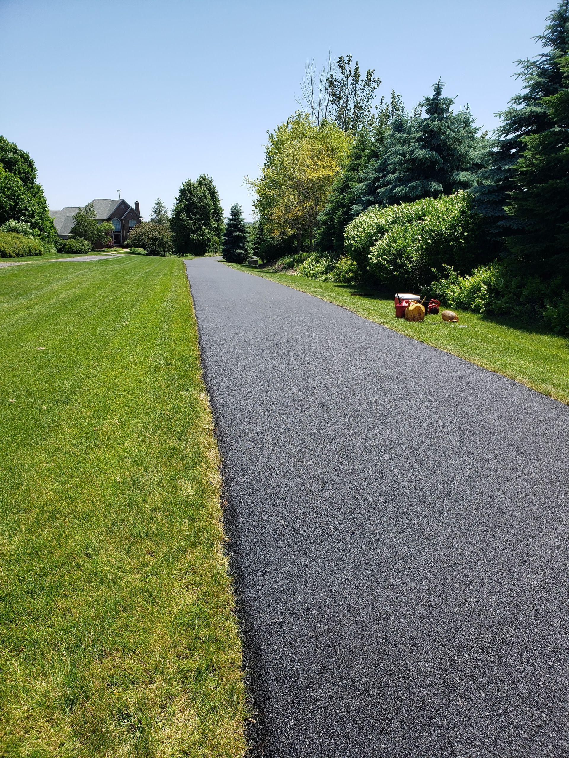 A straight asphalt path stretches through a grassy lawn toward trees and a distant house under a bright blue sky.