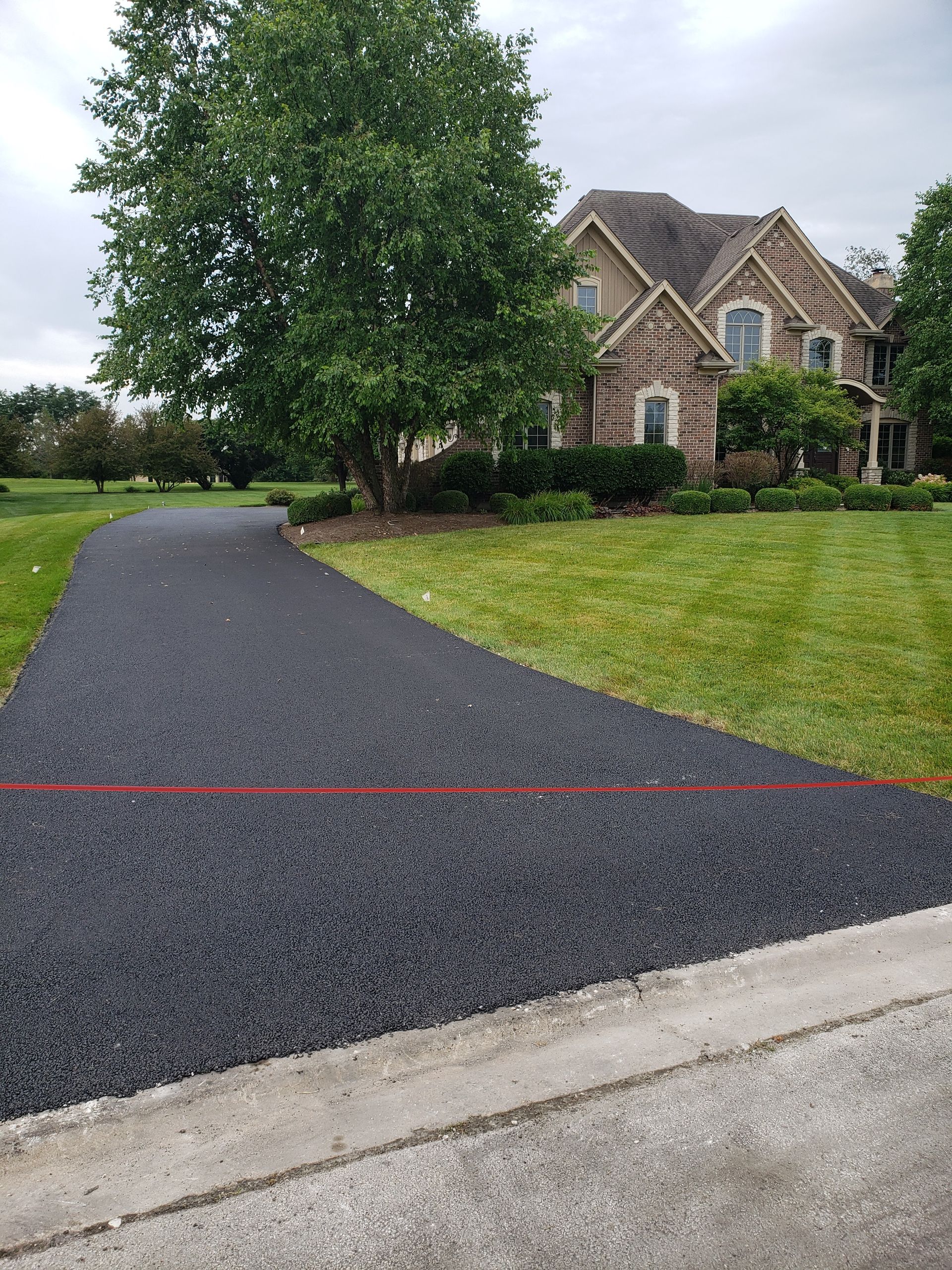 A newly paved black asphalt driveway leads to a large brick suburban home with a green lawn and trees.