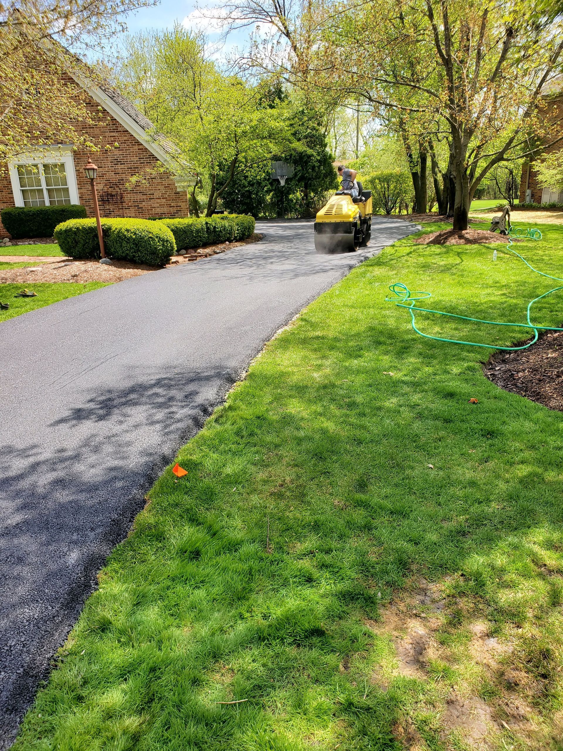 A yellow road roller compacts a fresh gravel driveway leading to a brick house on a sunny day.