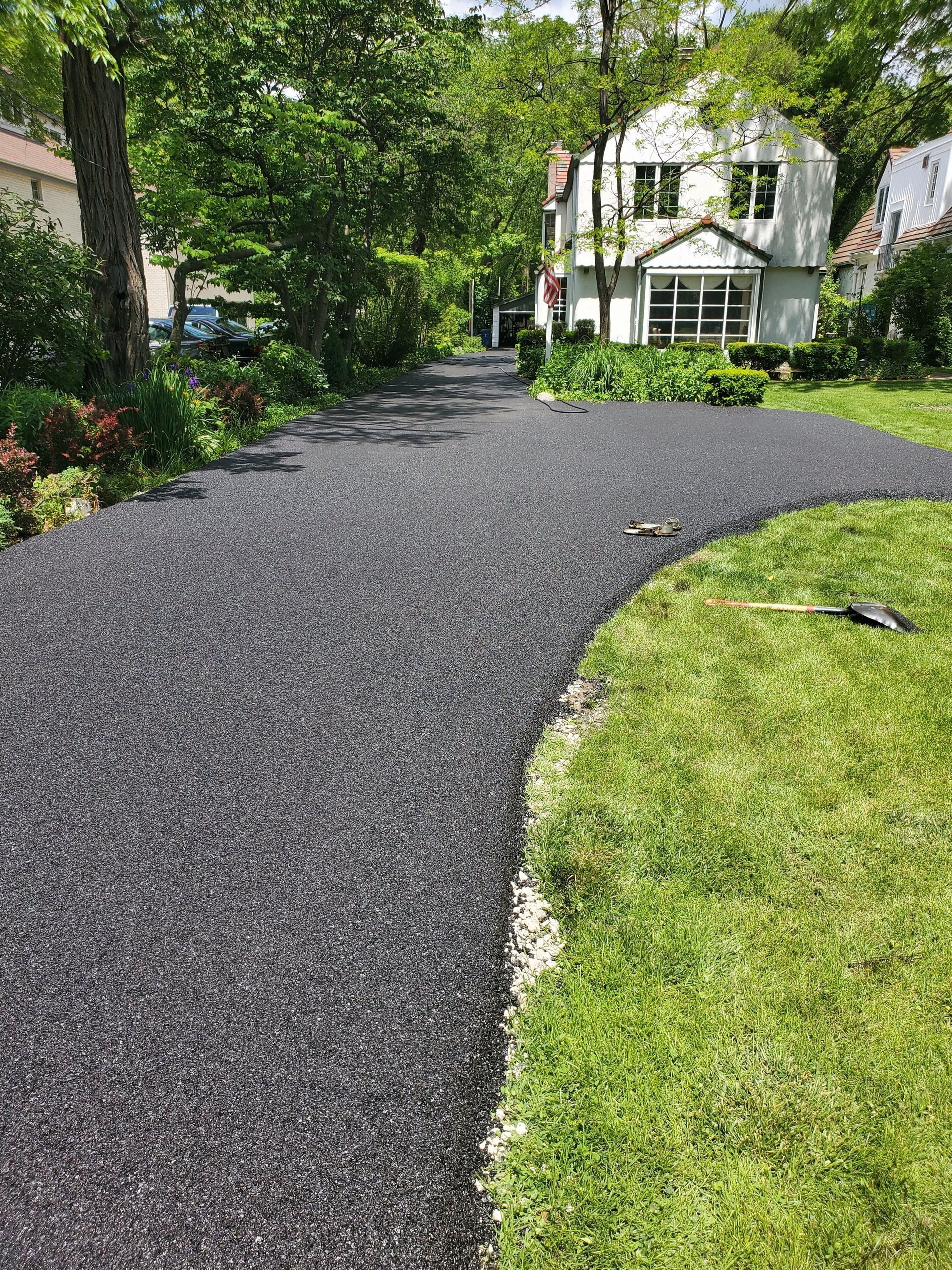 A freshly paved, dark gray gravel driveway curves through a green, grassy front yard toward a house.