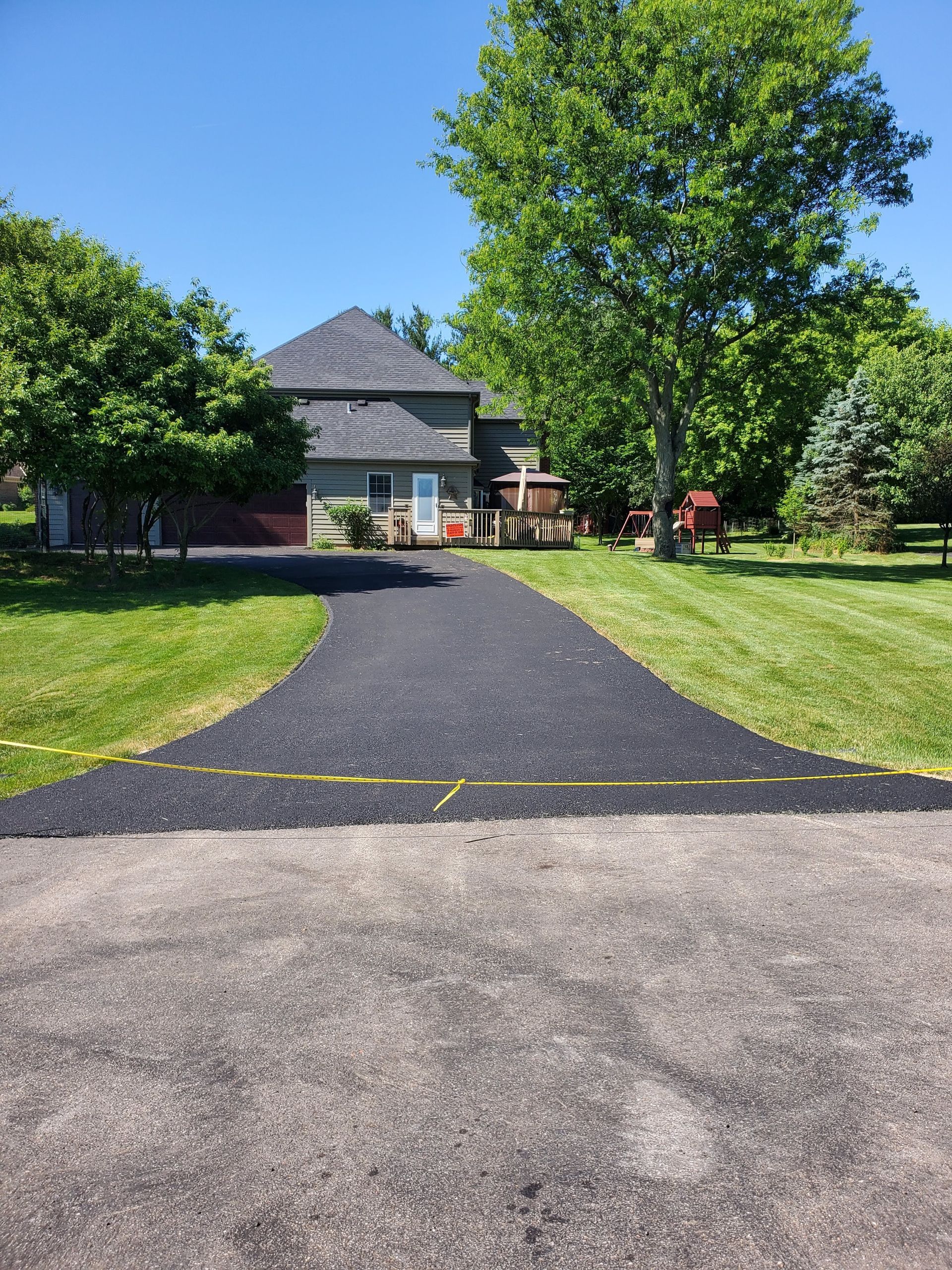 Freshly paved asphalt driveway leading to a house surrounded by green trees and lawn on a sunny day.