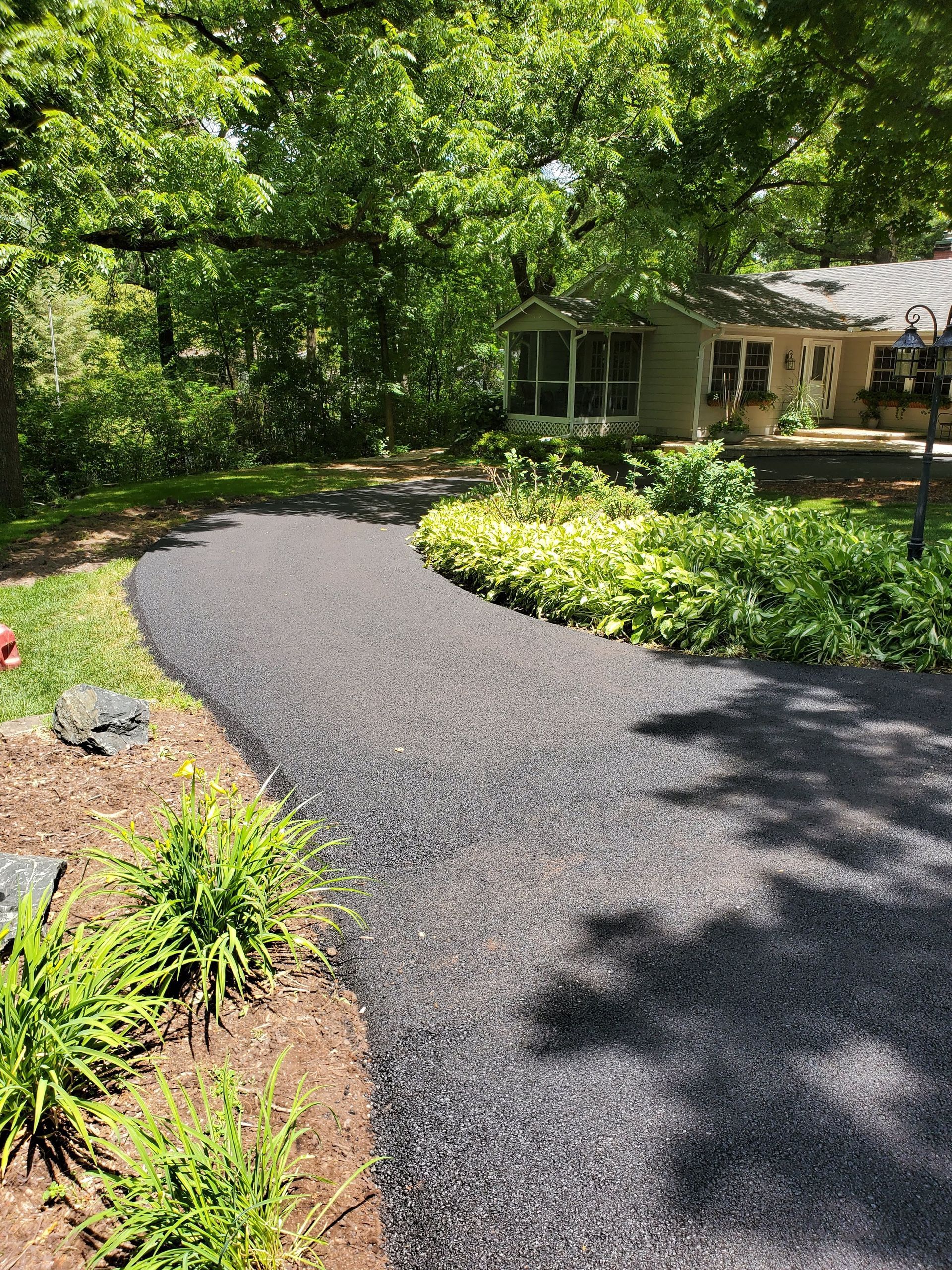 A dark, coarse-textured garden path winds through a lush, sunny yard toward a light-colored house with a screen porch.