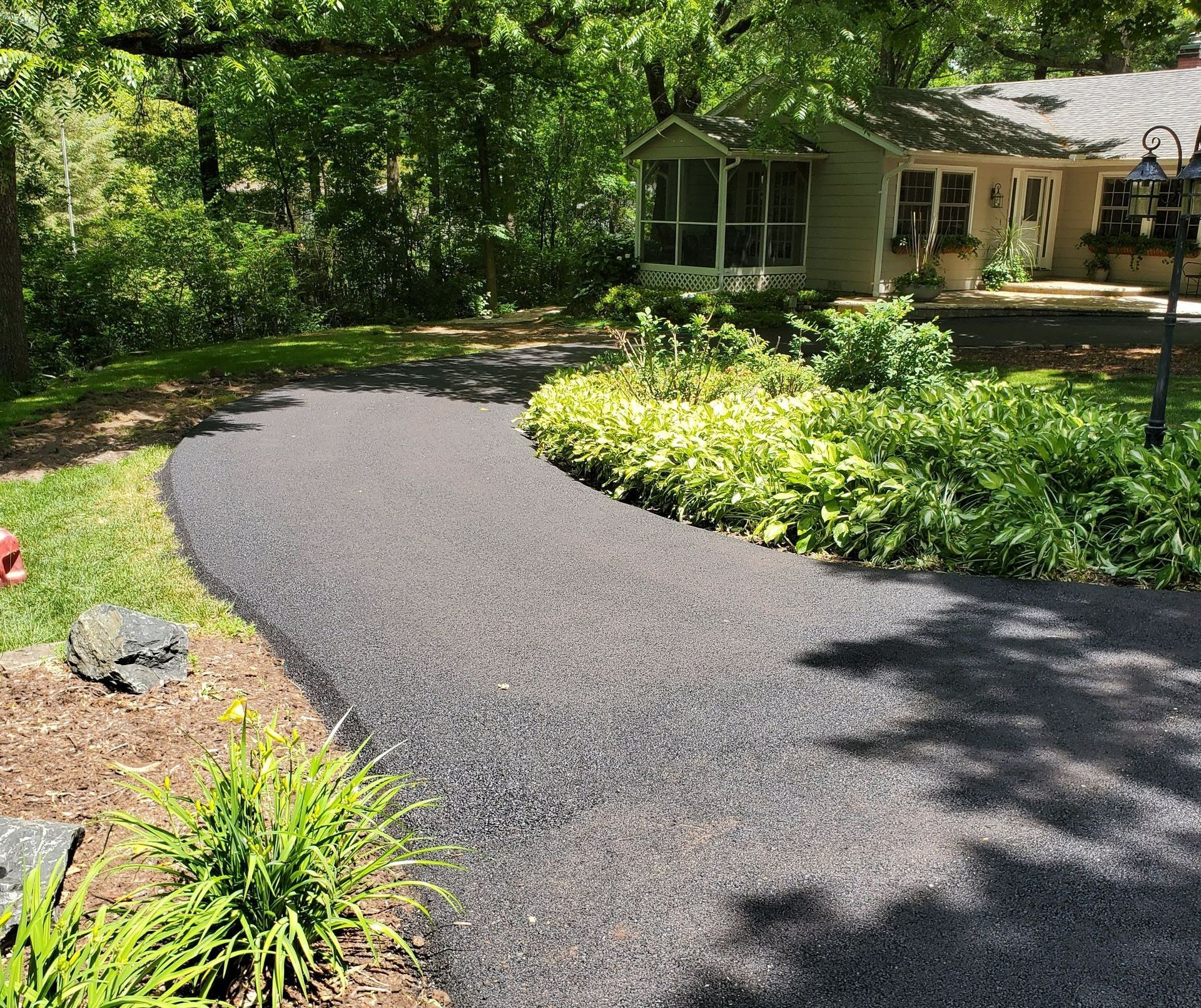 A newly paved asphalt driveway curves through a lush green yard towards a house with a screened porch in the background.