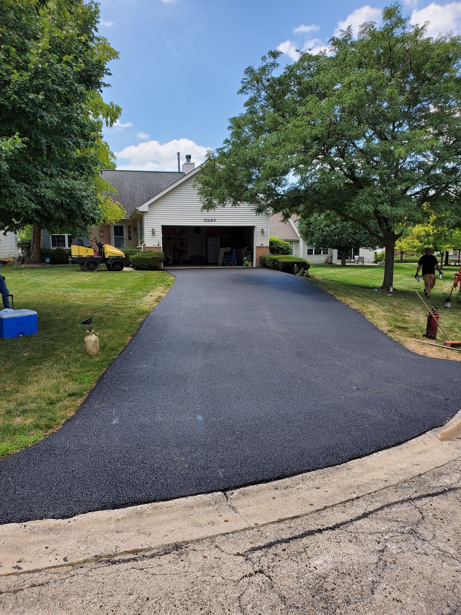 A newly paved, dark asphalt driveway leading to a suburban home garage on a sunny day.