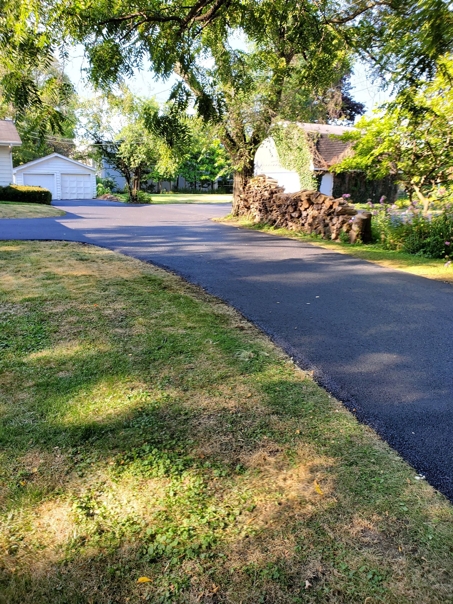 A paved driveway leads toward a white house with a garage and a large woodpile beneath trees on a sunny day.