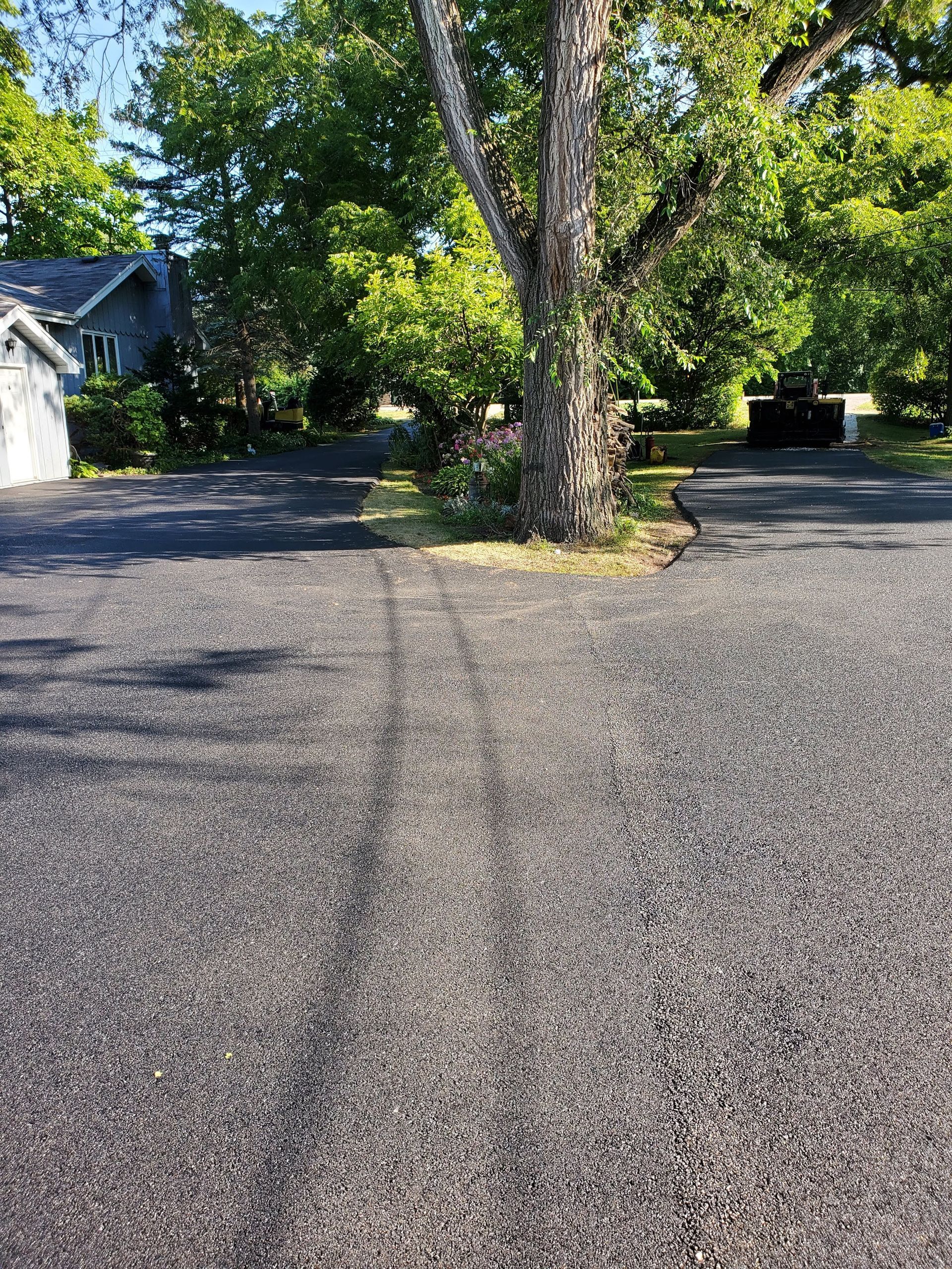 A gravel driveway splits into two paths around a large tree in front of a house, with tire tracks visible on the ground.