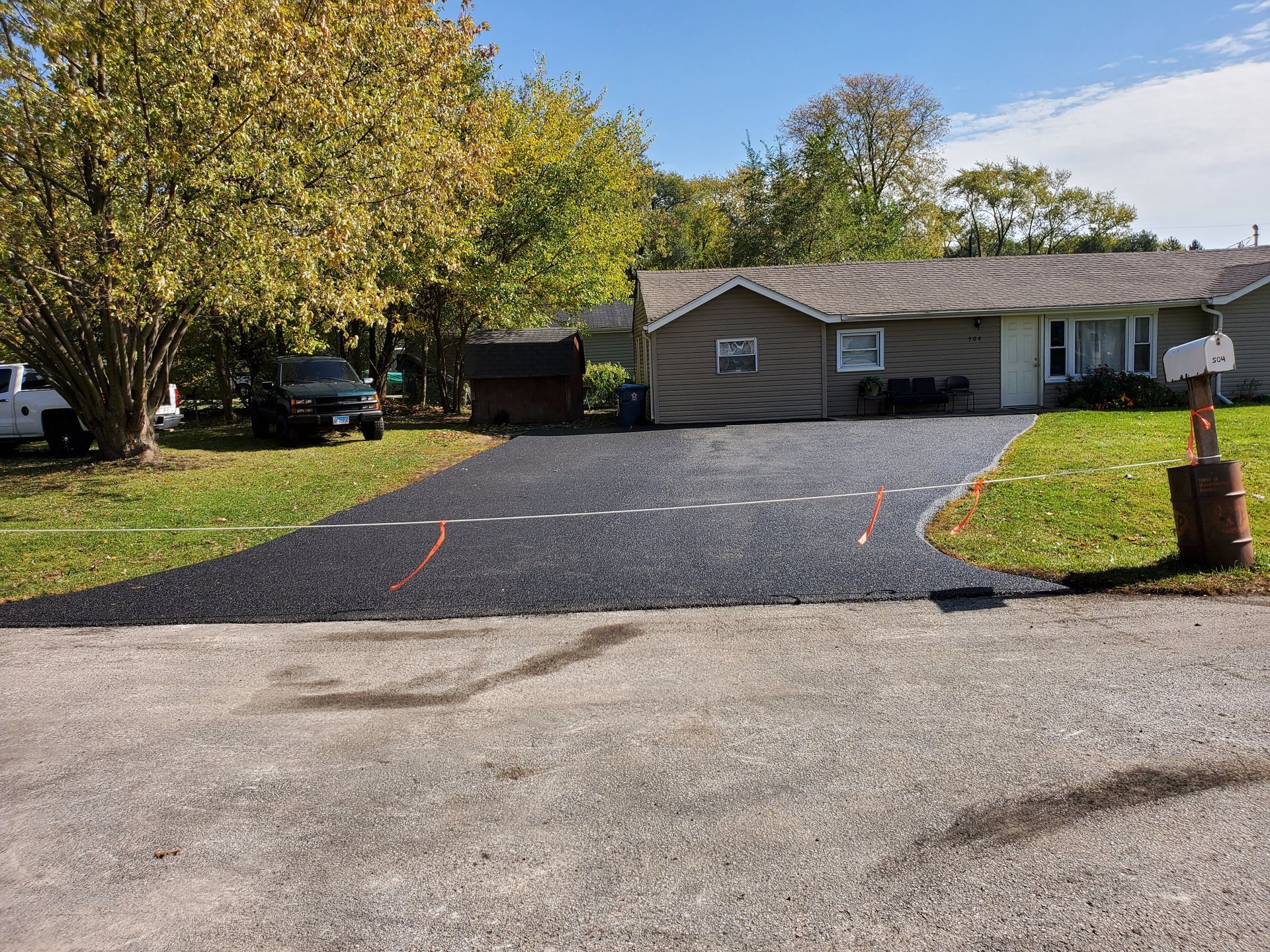 A newly paved black asphalt driveway leads to a ranch-style house, set against a backdrop of trees with autumn foliage.