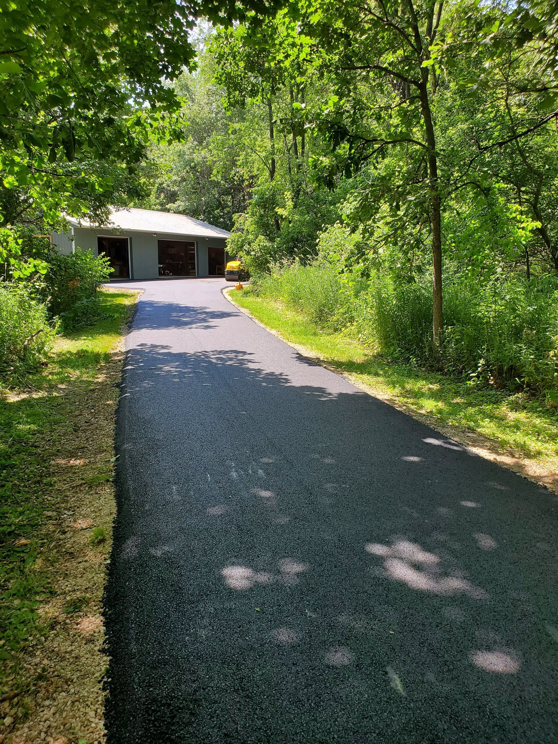 A freshly paved, dark asphalt driveway leads through a wooded area to a detached garage under bright, sunny conditions.