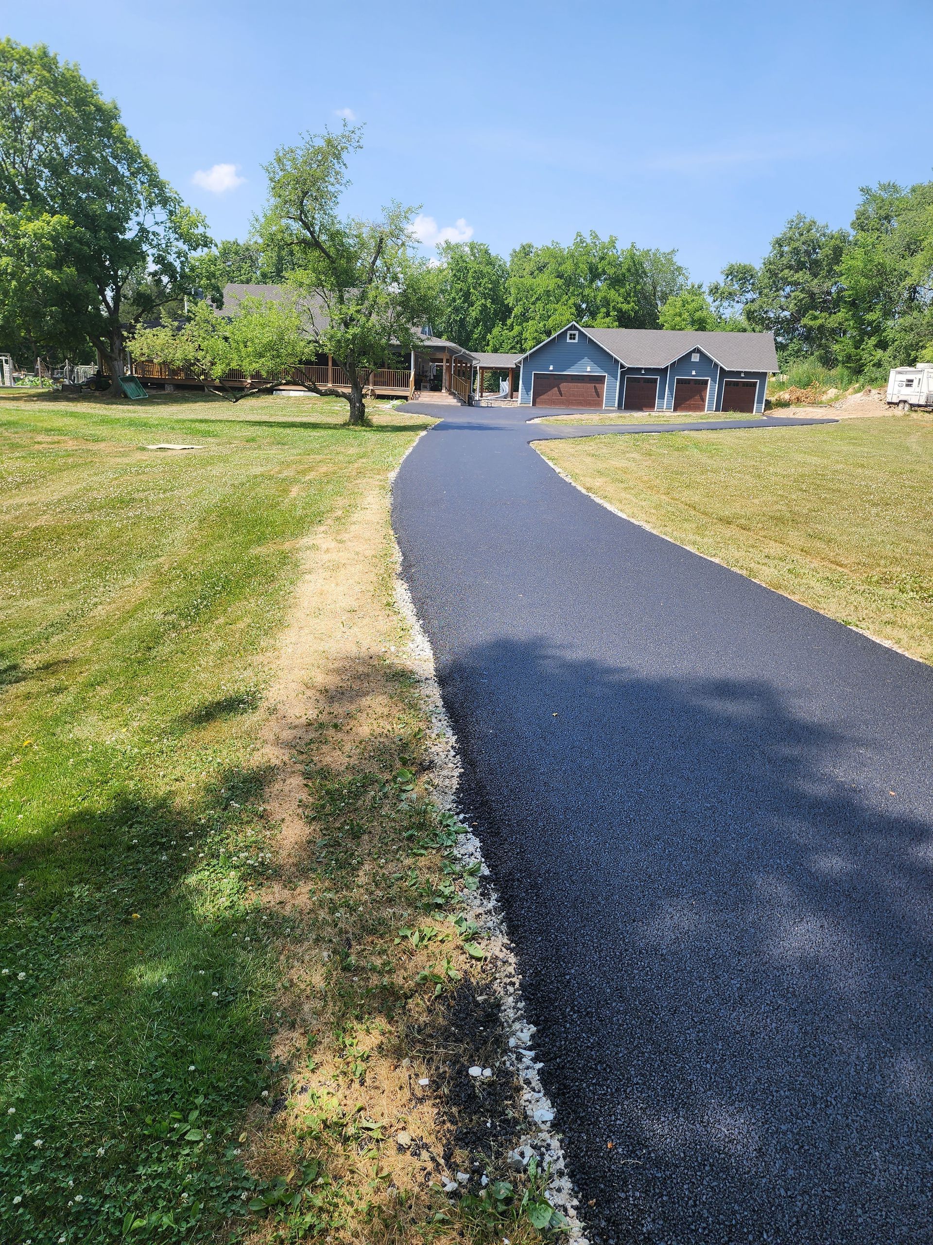 A freshly paved, dark asphalt driveway leads through a grassy yard toward a house with a three-car garage.
