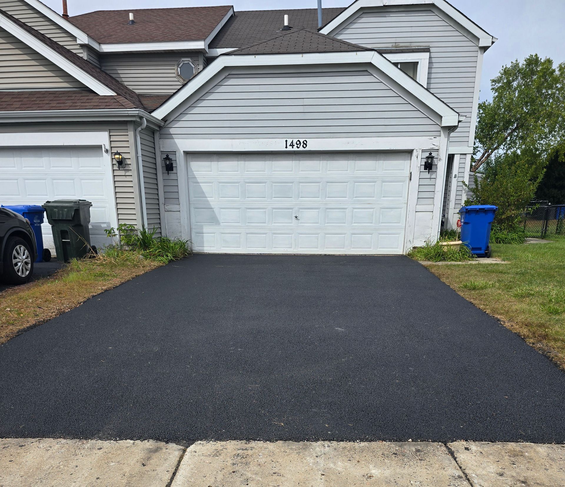 A newly paved dark asphalt driveway leads to a suburban garage with light-colored siding and a white door.