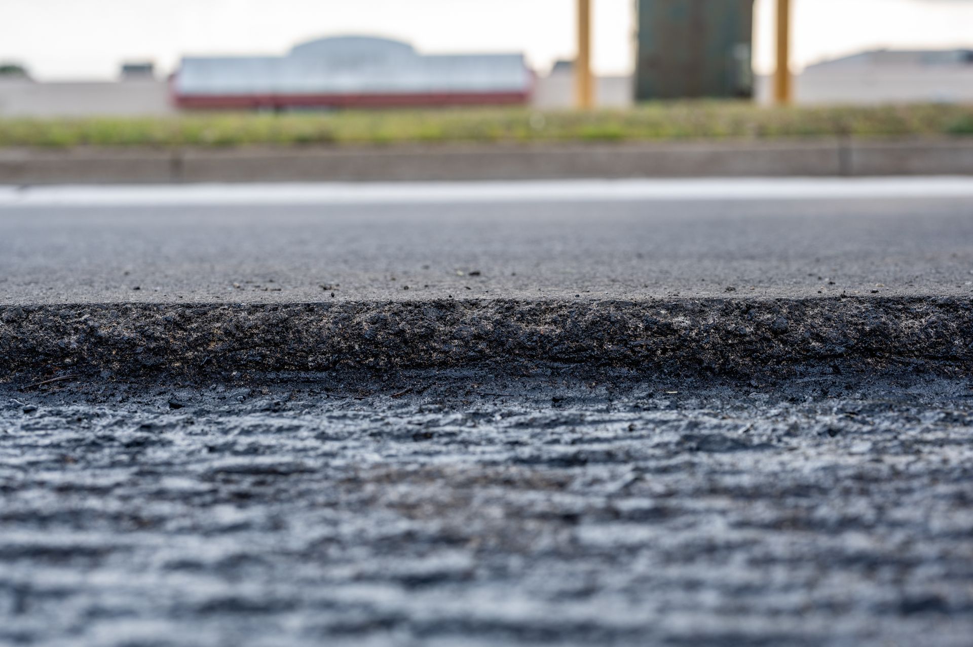 A close-up view of an asphalt road surface with a visible ledge between a freshly paved layer and the rougher base layer.