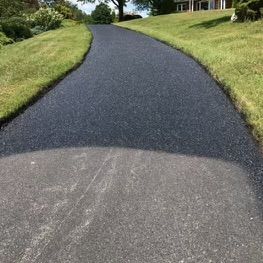 A freshly paved, dark asphalt driveway leads uphill between grassy lawns toward a house in the distance.