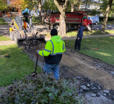 A crew with a skid-steer and a red dump truck removes old pavement from a driveway in a residential neighborhood.