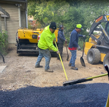 Three construction workers use shovels and a rake to spread fresh asphalt in front of a residential home.