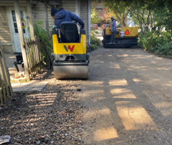 Two workers use yellow Wacker Neuson rollers to compact gravel on a driveway next to a wooden building.