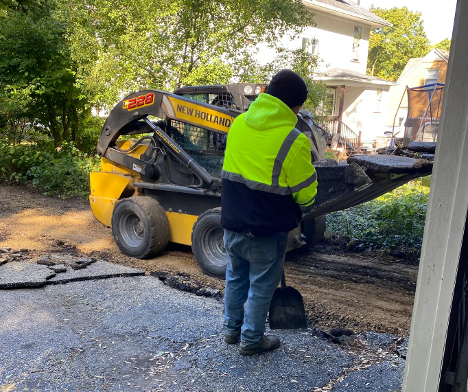 A person wearing a high-visibility jacket uses a shovel near a yellow skid-steer loader at a driveway construction site.