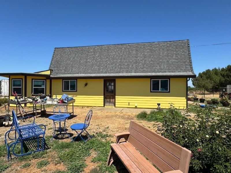 Yellow house with gray roof, blue chairs, and wooden bench on a sunny day.