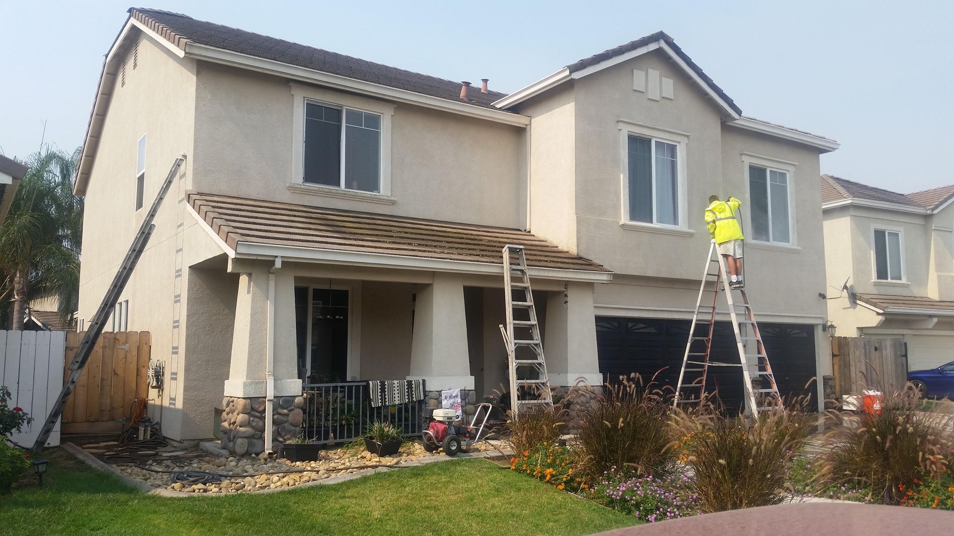 Two-story beige house with workers on ladders; green lawn and bushes.