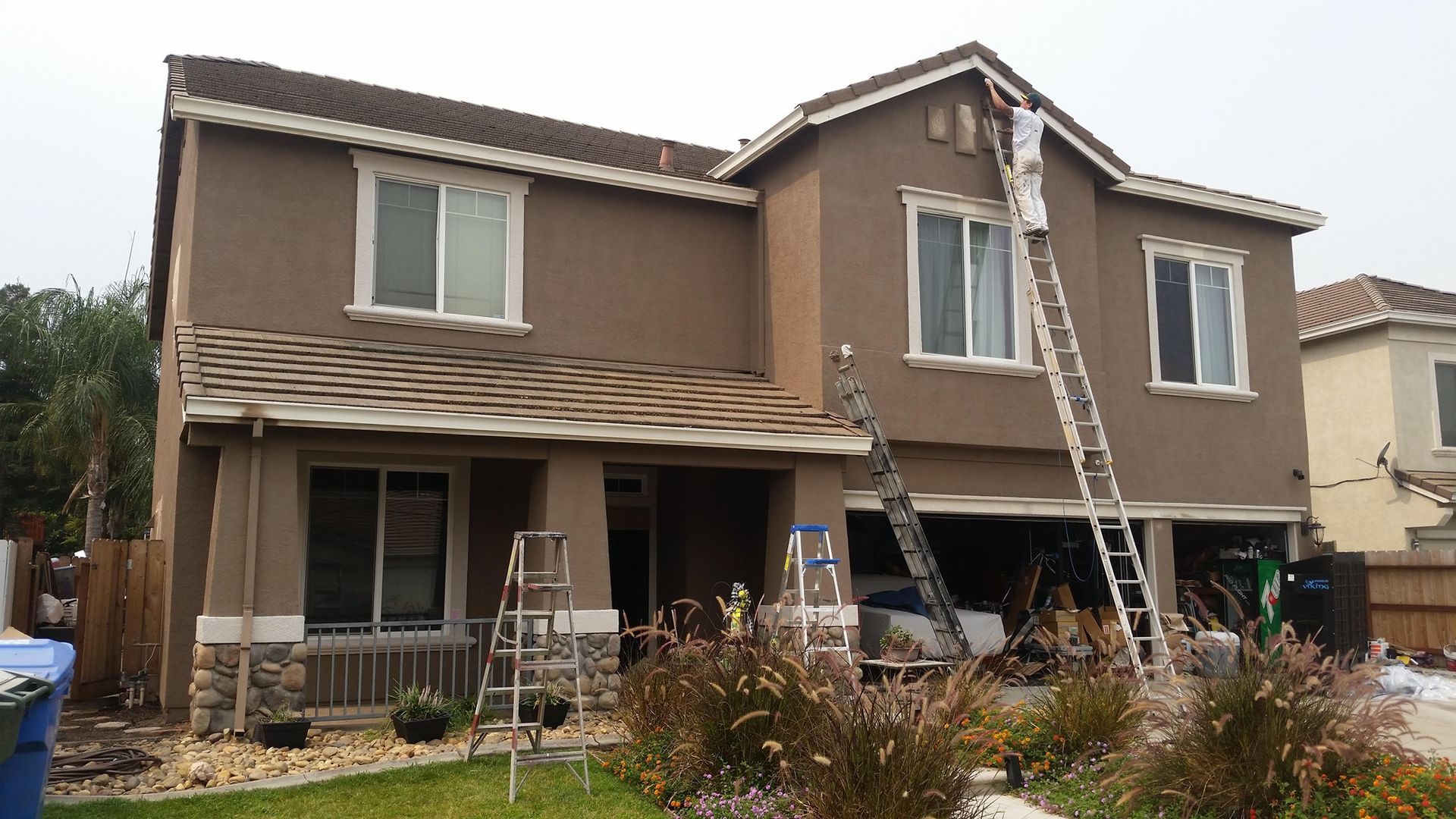 Two-story brown house with a person on a ladder painting the upper exterior, with bushes and green grass in front.