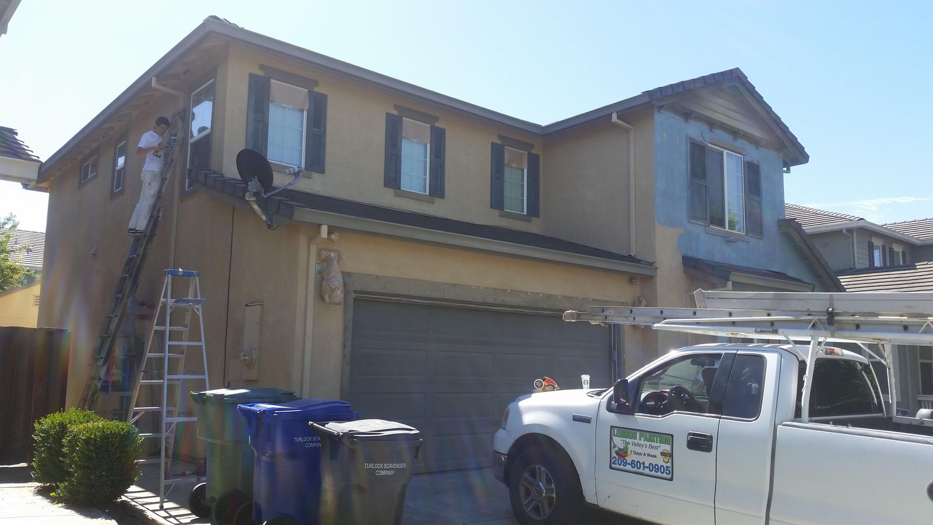 Workers painting a two-story house, light yellow exterior, blue accents, ladder, truck, and trash cans in front.