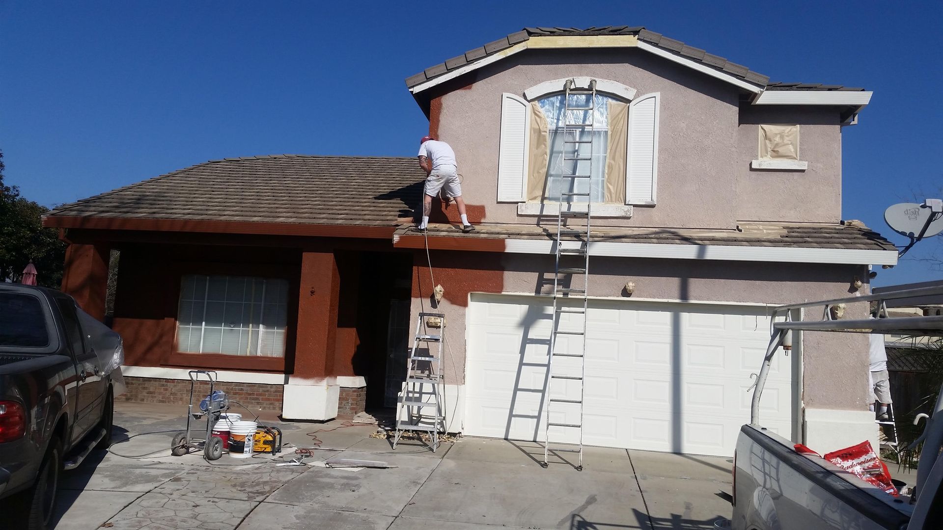 Person painting a two-story house exterior brown and beige with ladders on the driveway.