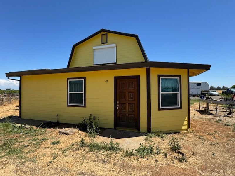 Yellow barn-shaped building with brown trim, door, and a white roll-down shutter on a clear, sunny day.