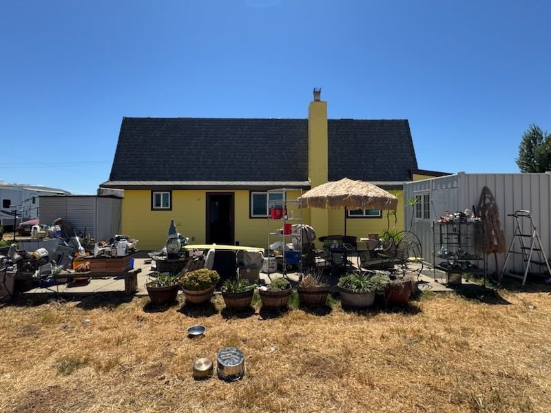 Yellow house with black roof, cluttered yard, plants, and blue sky.