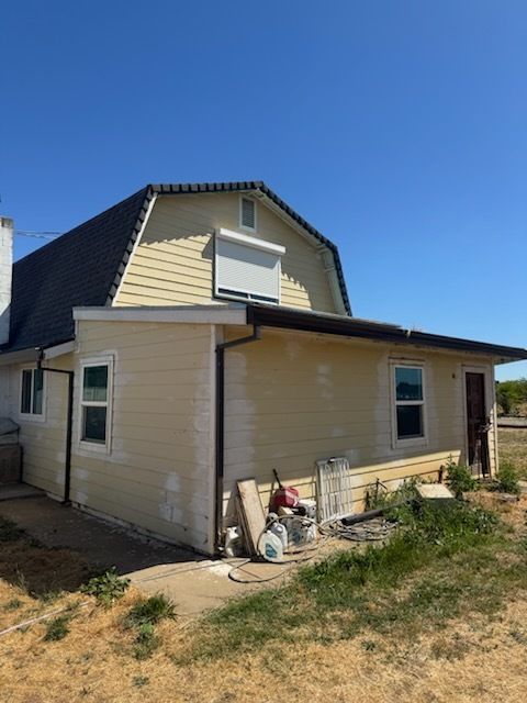 Side view of a two-story beige house under clear blue sky, showing unfinished paint patches and construction materials.