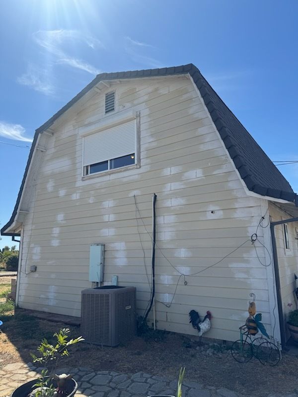 Beige barn-style building with white patches, air conditioning unit, black roof, under a blue sky.