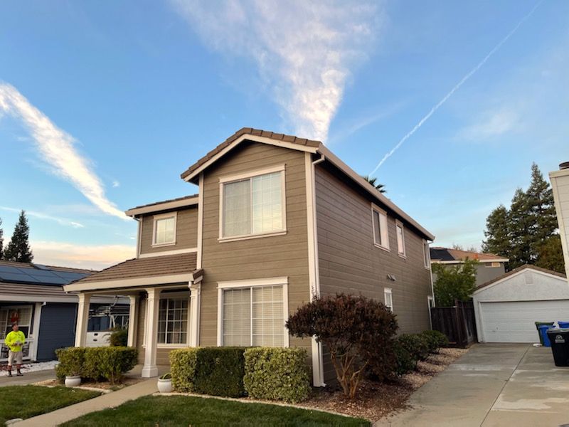 Two-story brown house with white trim, blue sky, and cloud streaks; driveway to a garage.