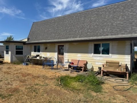 Back of a one-story house with a gray roof and tan siding, surrounded by dry grass and a blue sky.