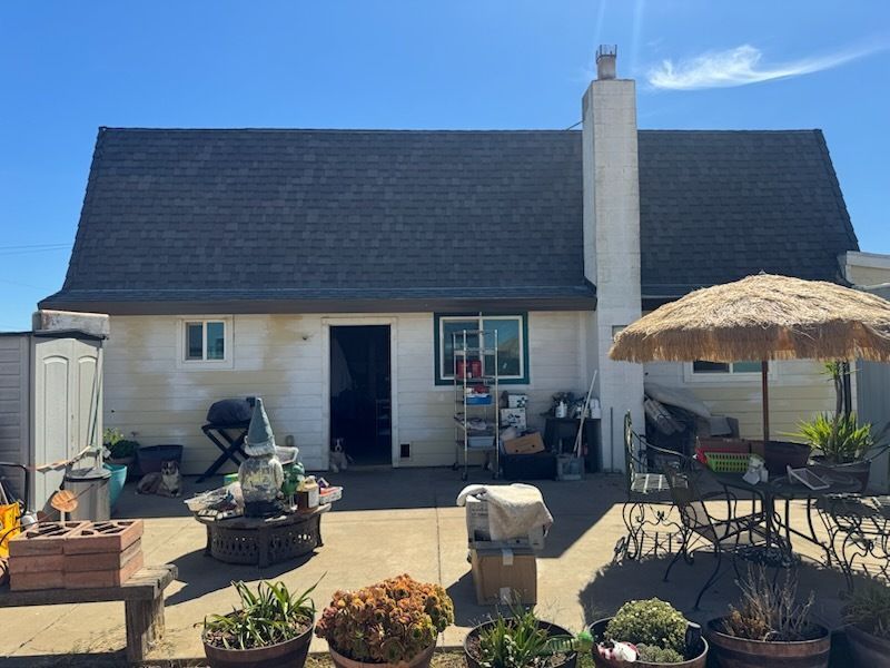 Backyard view of a white house with a dark roof, chimney, and patio. Plants, furniture, and a blue sky are present.