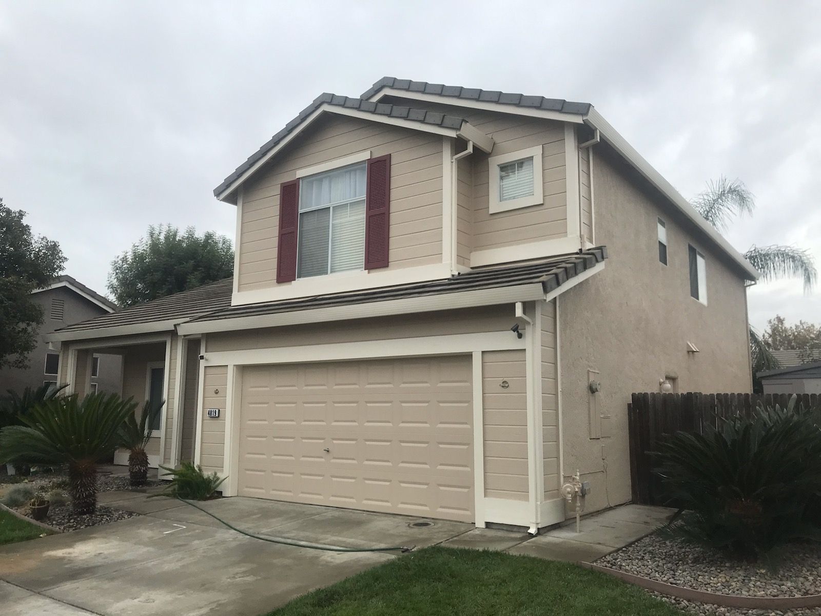 Two-story beige house with a tan garage door, red shutters, and a cloudy sky.