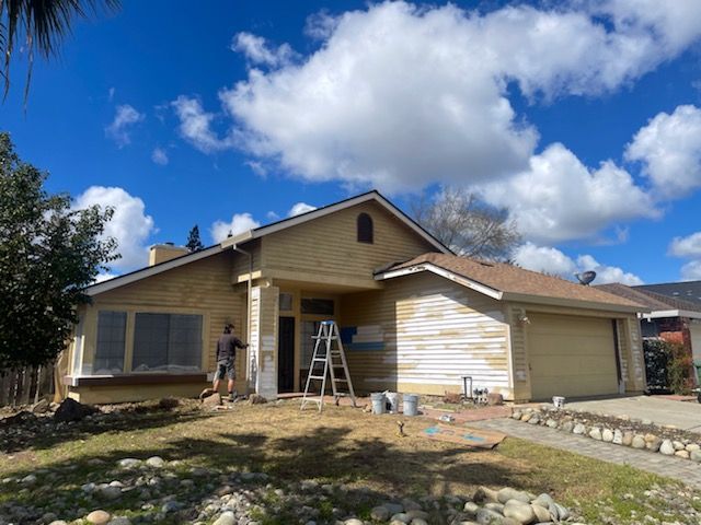 House being painted; light yellow siding partially primed, ladder present, person painting on porch. Blue sky.