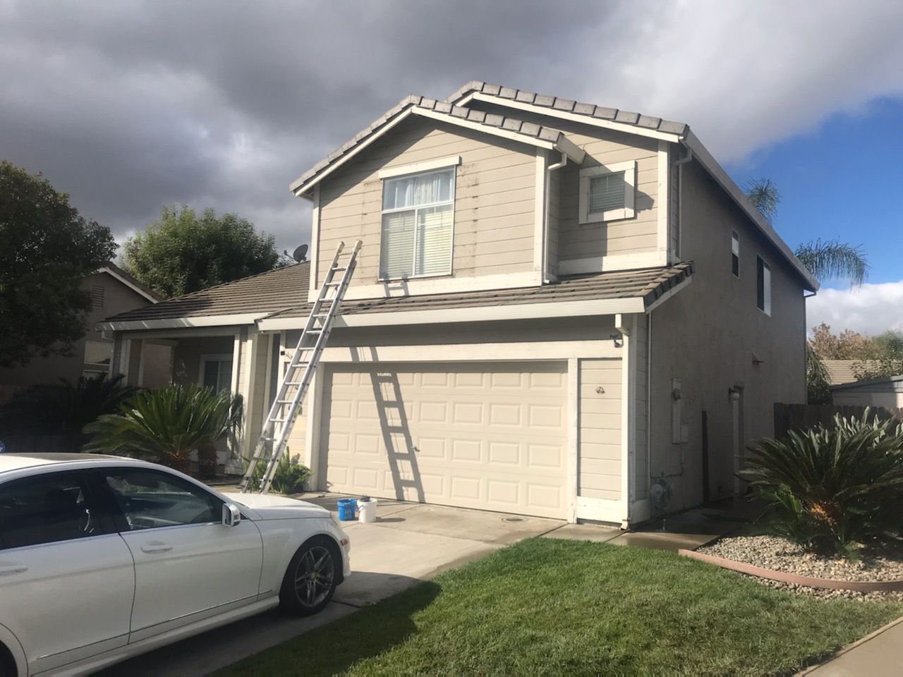 A two-story house with a ladder leaning against it, car in front, cloudy sky.
