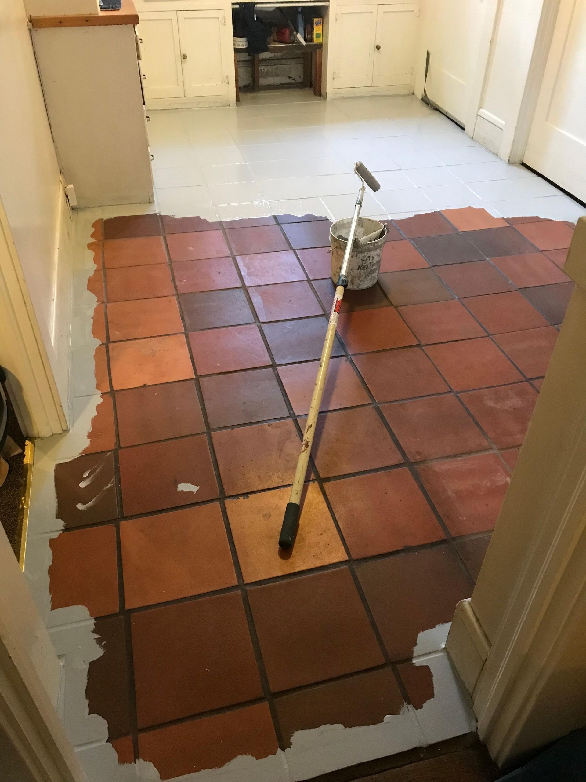 Brown tile floor being painted with gray around the edges, roller and paint can on tiles.