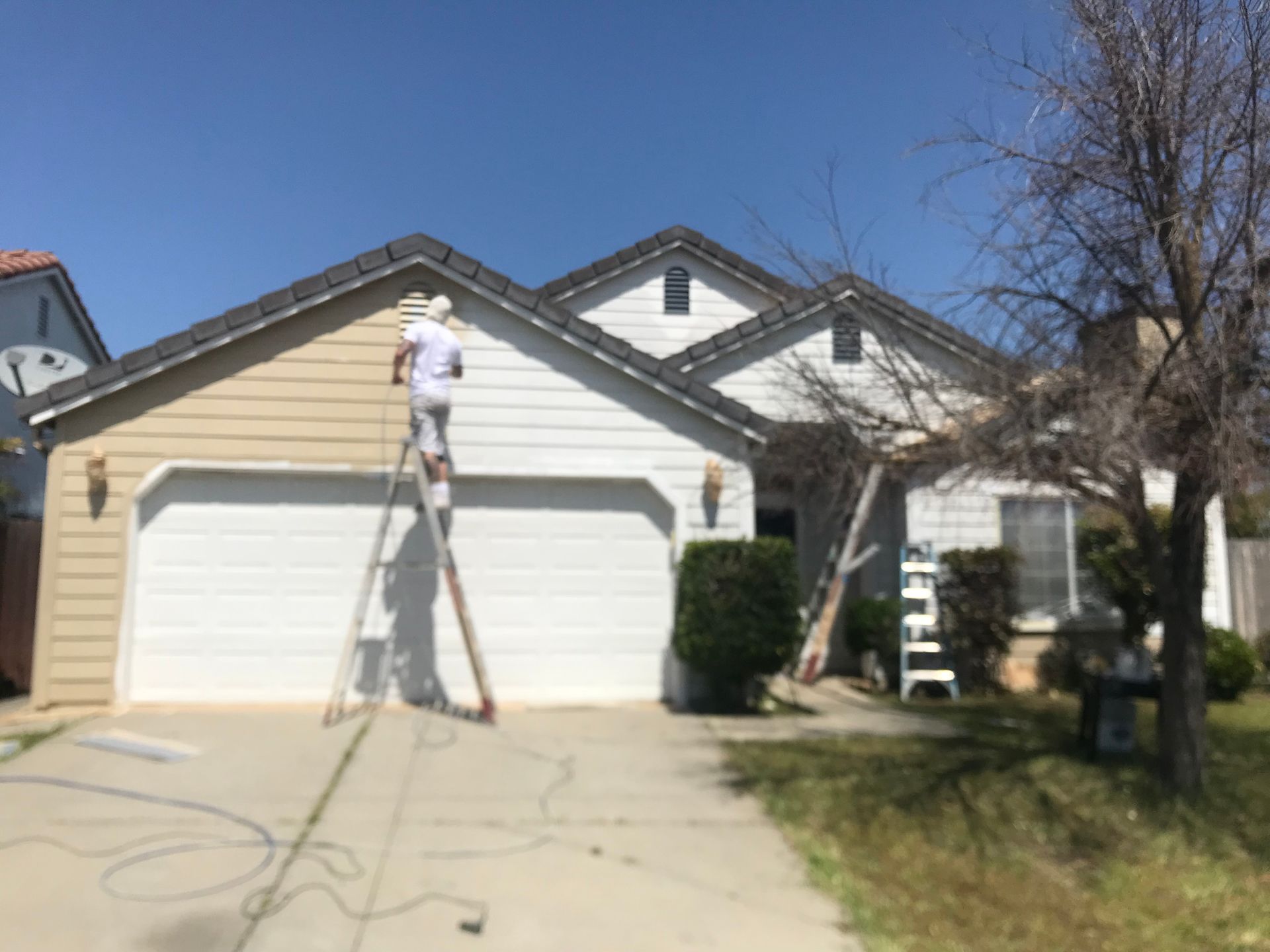 Person painting a beige house, standing on a ladder. White garage door, blue sky, and a tree are visible.