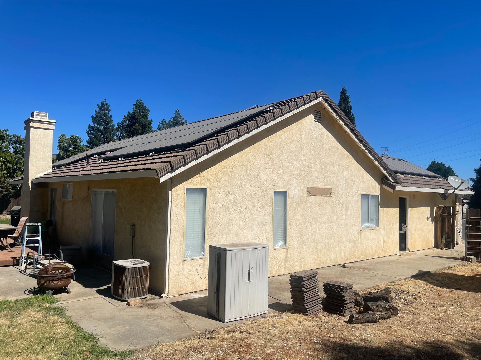 Beige stucco house with dark tiled roof, chimney, and backyard patio on a sunny day.