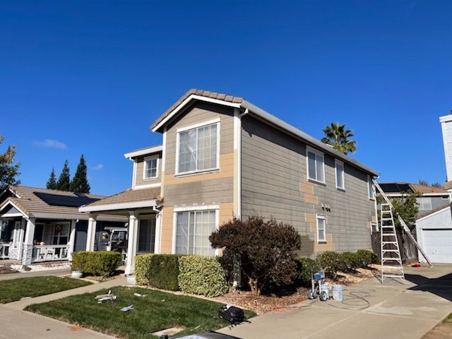 Two-story house being painted. Tan siding with sections painted gray, blue sky, and ladder propped up.