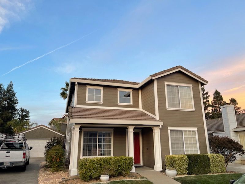 Two-story house with olive green siding, white trim, and red front door under a sunset sky.