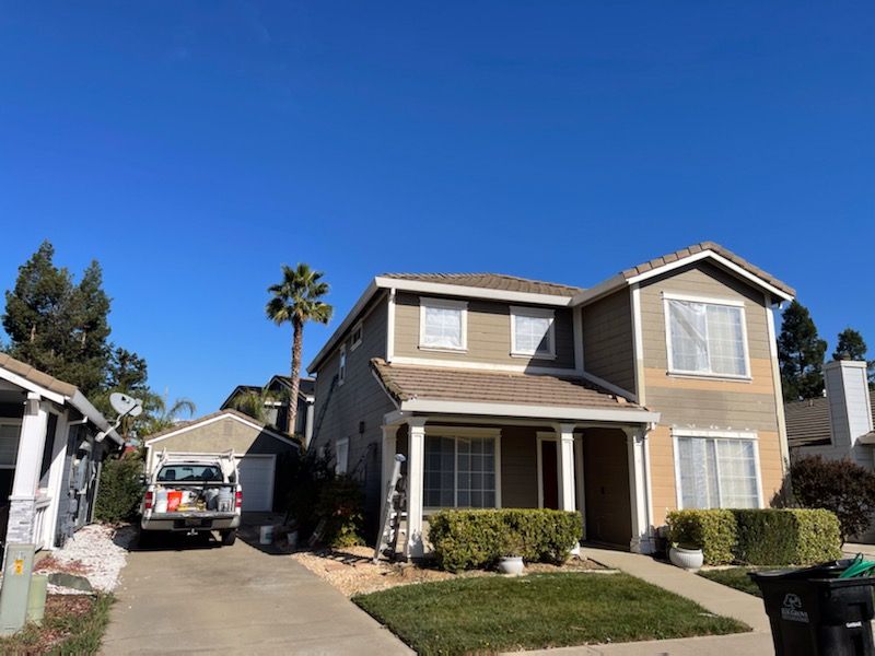 Two-story house with tan siding, green bushes, and a clear blue sky. A truck is parked in the driveway.
