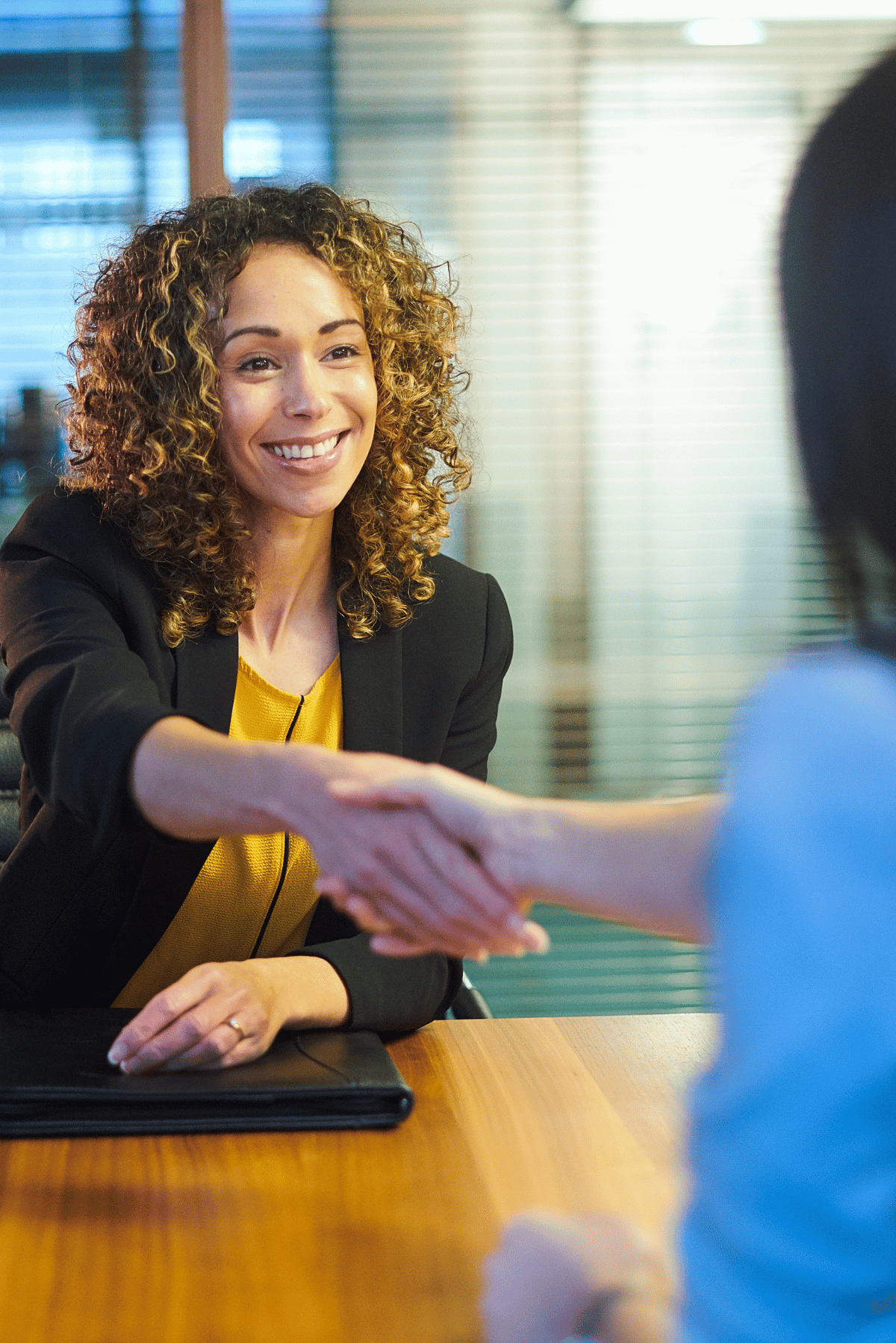 Two people shake hands across a table in a bright office setting.