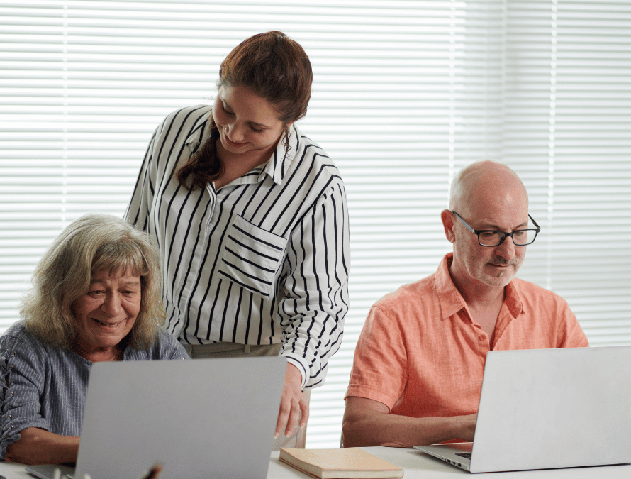 Three people using laptops at a bright table, with one woman standing and helping.