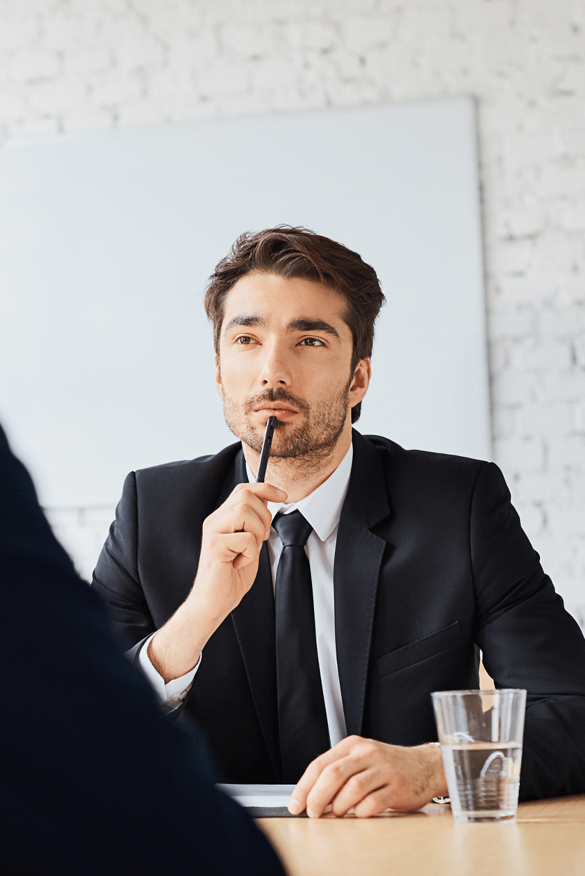 Businessman in a suit thinking at a desk, holding a pen, with a glass of water nearby.