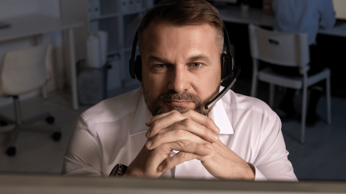 Man in white shirt wearing a headset, resting clasped hands under his chin in an office setting