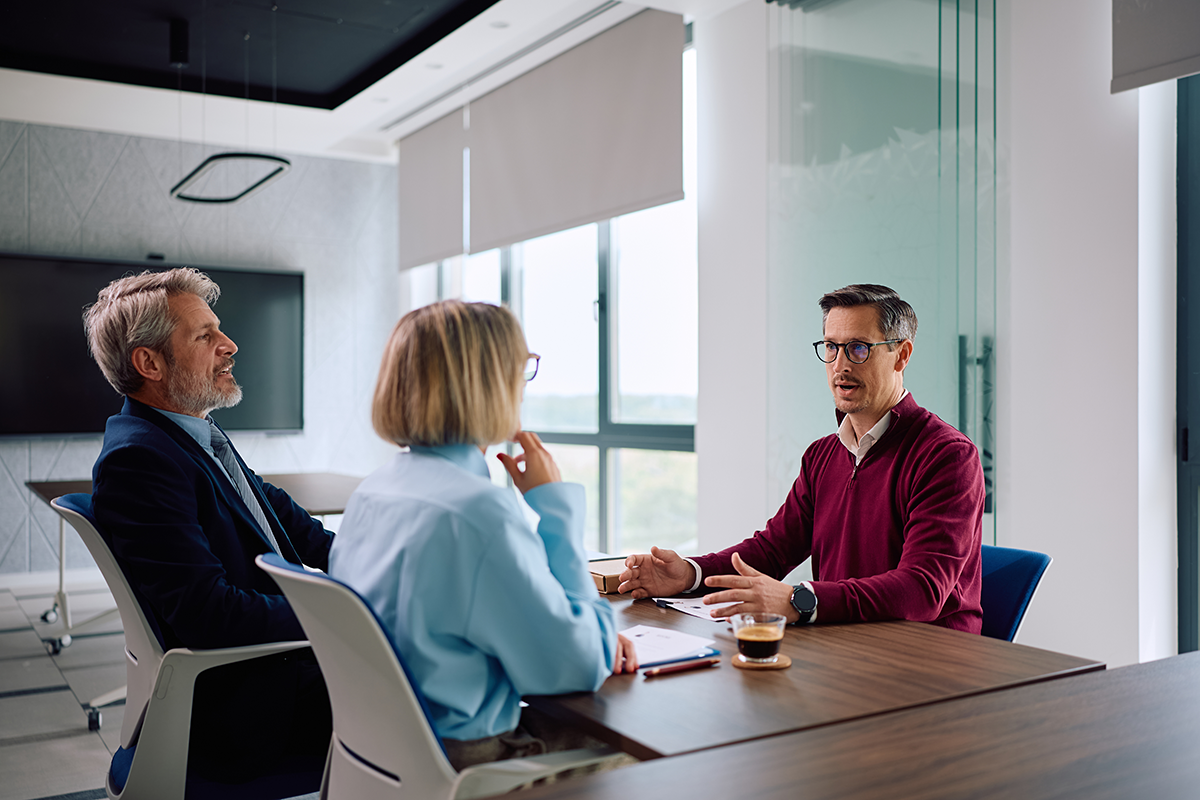 Three people in a modern office meeting around a conference table, talking with laptops and coffee cups.
