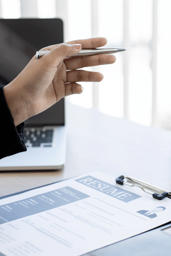 Hand holding a pen over a clipboard with paperwork beside a laptop on a desk