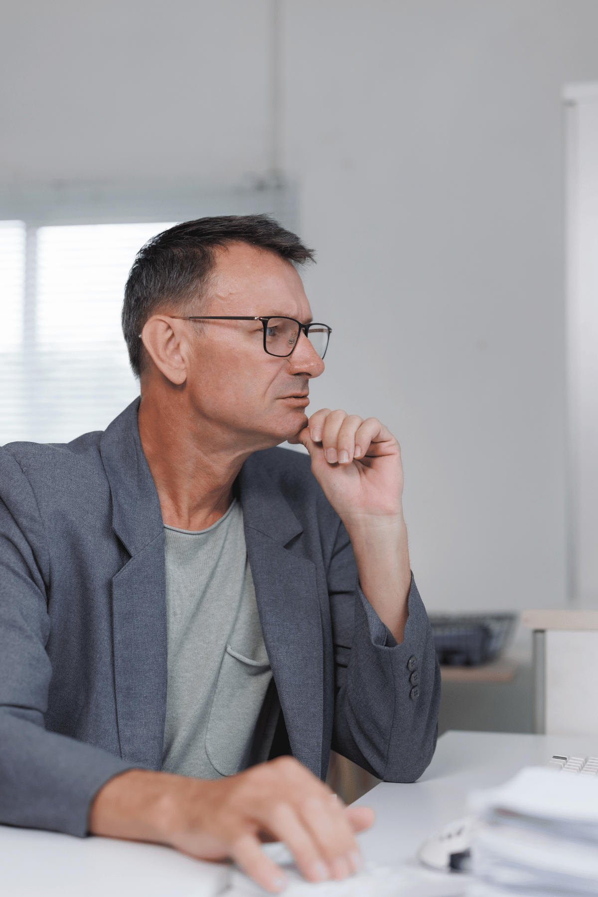 Man in glasses in a gray blazer sits at a desk, resting his chin and looking thoughtfully to the side.