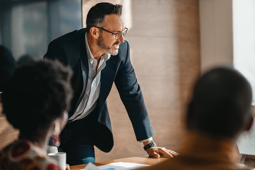 Businessman leaning over a conference table, speaking to seated colleagues in a meeting room.