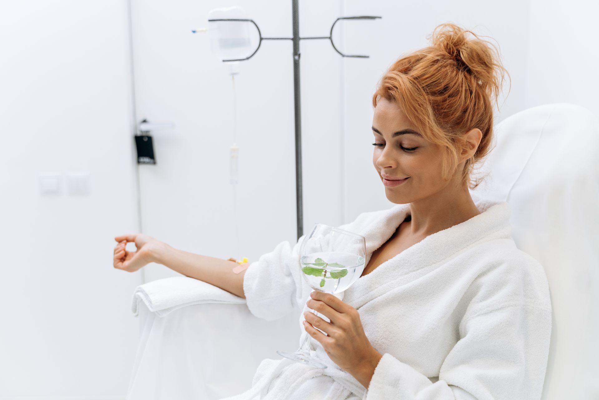 Woman in white robe receiving IV, smiling, holding a drink, in a bright room.